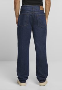 Mies, joka pukeutunut tummansinisiin suoralahkeisiin farkkuihin ja valkoisiin tennareihin, seisoo selkä kameraan päin yksiväristä harmaata taustaa vasten.