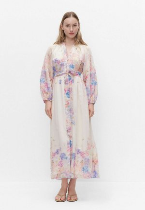 Kvinde iført en lang, lys blomsterkjole med lange ærmer og taljebånd, stående mod en ensfarvet hvid baggrund, vendt fremad.