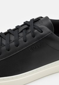 Svart lädersneaker med texturerad yta, kontrasterande vit sula och platta svarta sn ören. Har präglad "BOSS"-logotyp på sidan.