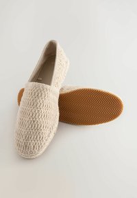 Zapatos de lona beiges tipo slip-on con un upper texturizado y suela de goma con un patrón de espiga, diseñados para la comodidad y el uso casual.