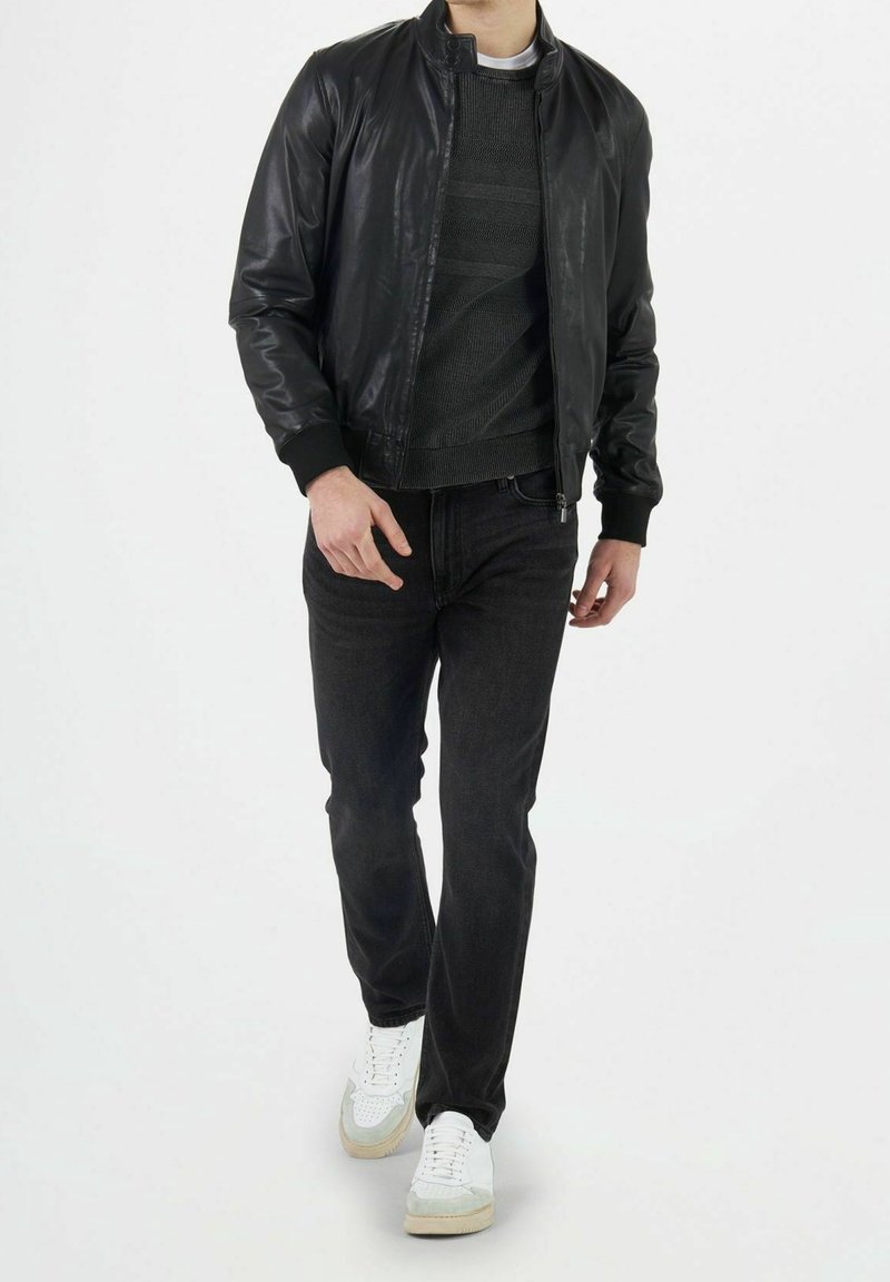 Giubbotto bomber in pelle nera sopra un maglione grigio a righe, abbinato a jeans scuri e sneaker bianche con dettagli grigi.