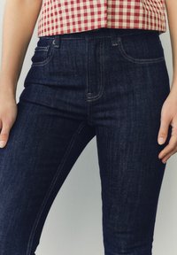 Tmavé denimové džínsy so slim fit strihom, s kontrastným prešívaním a prednými vreckami. Materiál má textúrovaný, mierne opotrebovaný vzhľad.