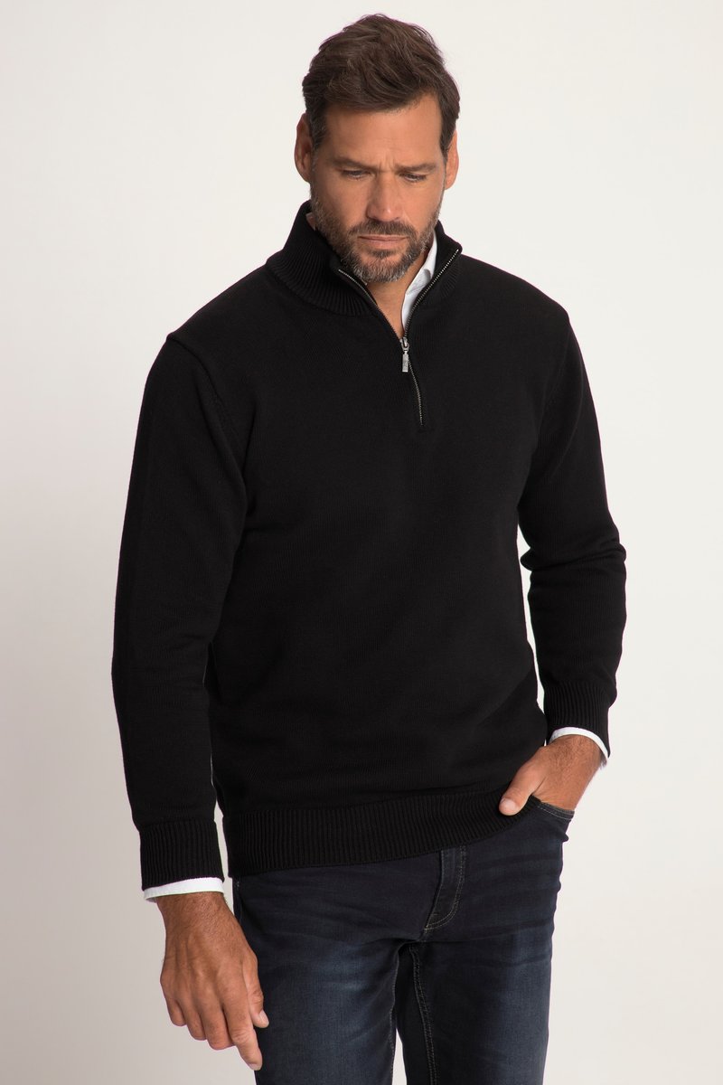 Mann trägt schwarzen Pullover mit halbem Reißverschluss über weißem Hemd, mit einer Hand in der Tasche und schaut nach unten vor neutralem Hintergrund.