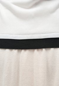 Balta blūze ar strukturētu virsmu un melnu elastīgu jostasvietu. Apakšējā daļa izgatavota no maiga, ribota bēša auduma.