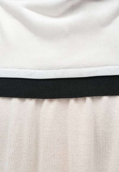 Haut blanc avec une finition texturée, doté d'une ceinture élastique noire. La partie inférieure est en tissu beige doux côtelé.