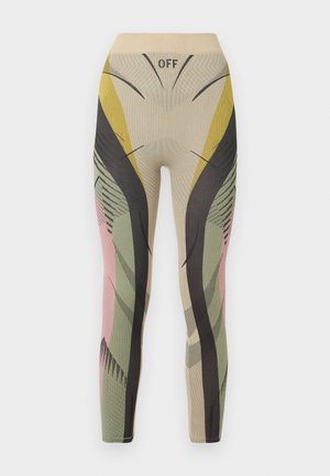 Kobieta stojąca z ręką na biodrze, ubrana w wielokolorowy, wzorzysty, dopasowany kombinezon oraz czarne sandały na wysokim obcasie na gładkim szarym tle.