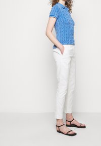 Modré krátké rukávové polo tričko s kabelovým pletením, bílé úzké kalhoty a černé sandály na pásky, které ukazují texturované pletení a čisté linie.