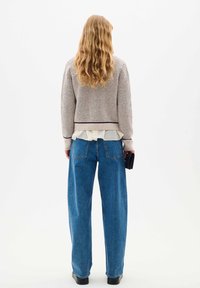 Crochet knit cardigan in beige and gray, layered over a light ruffled top; paired with high-waisted, loose blue jeans and black shoes.