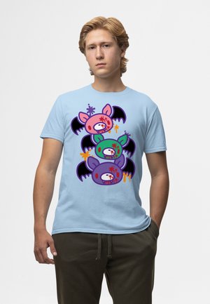 Ung mand iført lyseblå T-shirt med farverige tegneserie-flagermushoveder stablet lodret, med rødt sprøjt og sorte vinger, mod ensfarvet baggrund.