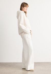 Woman standing sideways wearing an oversized cream hoodie, wide-legged white pants, and beige shoes against a plain white background.