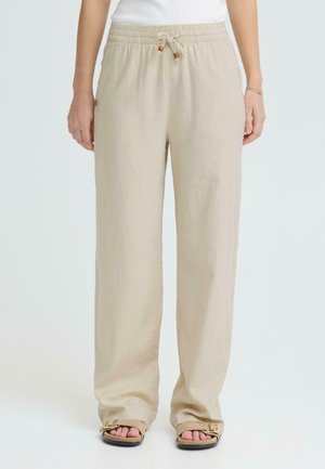 Person iført beige vide linbukser med snor i livet og brune åpne sandaler, stående mot en ensfarget bakgrunn.