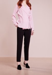 Vrouw in een lichtroze wikkelblouse met zijknopen, zwarte op maat gemaakte broek en zwarte slingback hakken, staande tegen een neutrale achtergrond.