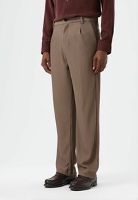 Pantalon beige plissé en tissu texturé, doté d'une boucle de ceinture et d'une fermeture à bouton, porté avec des mocassins marron foncé.