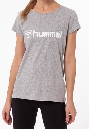 Kobieta ubrana w jasnoszarą koszulkę marki hummel® z krótkim rękawem oraz czarne spodnie na białym tle.