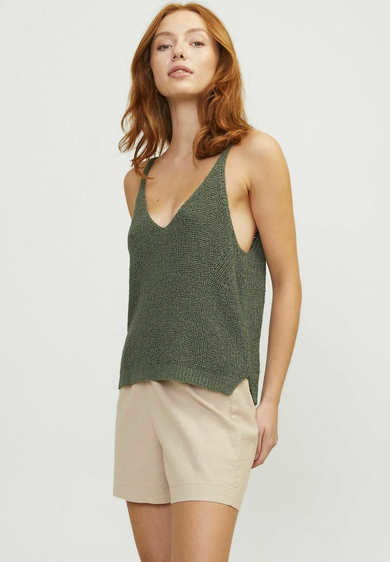 Top de punto verde con escote en V, aberturas laterales y tirantes anchos, combinado con pantalones cortos beige claro, presentado sobre un fondo neutro.