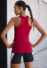 Rotes sportliches Tanktop mit Racerback-Design, kombiniert mit schwarzen Shorts mit weißen seitlichen Akzenten. Das Material wirkt glatt und enganliegend.