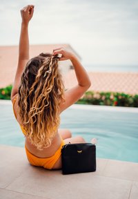 Žena v oranžových plavkách sedící vedle bazénu, hrající si s dlouhými kudrnatými vlasy, vedle ní černá taška, v pozadí obklopená dlaždicovou střechou a zelení.