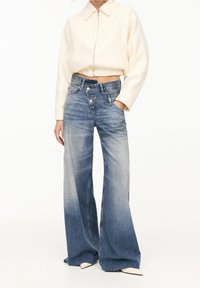 Jeans de mezclilla de pierna ancha en un lavado azul desvanecido, con una cintura alta, cierre de dos botones y detalles de desgaste sutil. Acompañados de una chaqueta corta color crema.