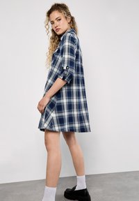 Niebieska i biała sukienka w kratę z podwiniętymi rękawami, guzikami oraz rozszerzanym krojem, wykonana z miękkiego materiału. Na nogach czarne buty na platformie.