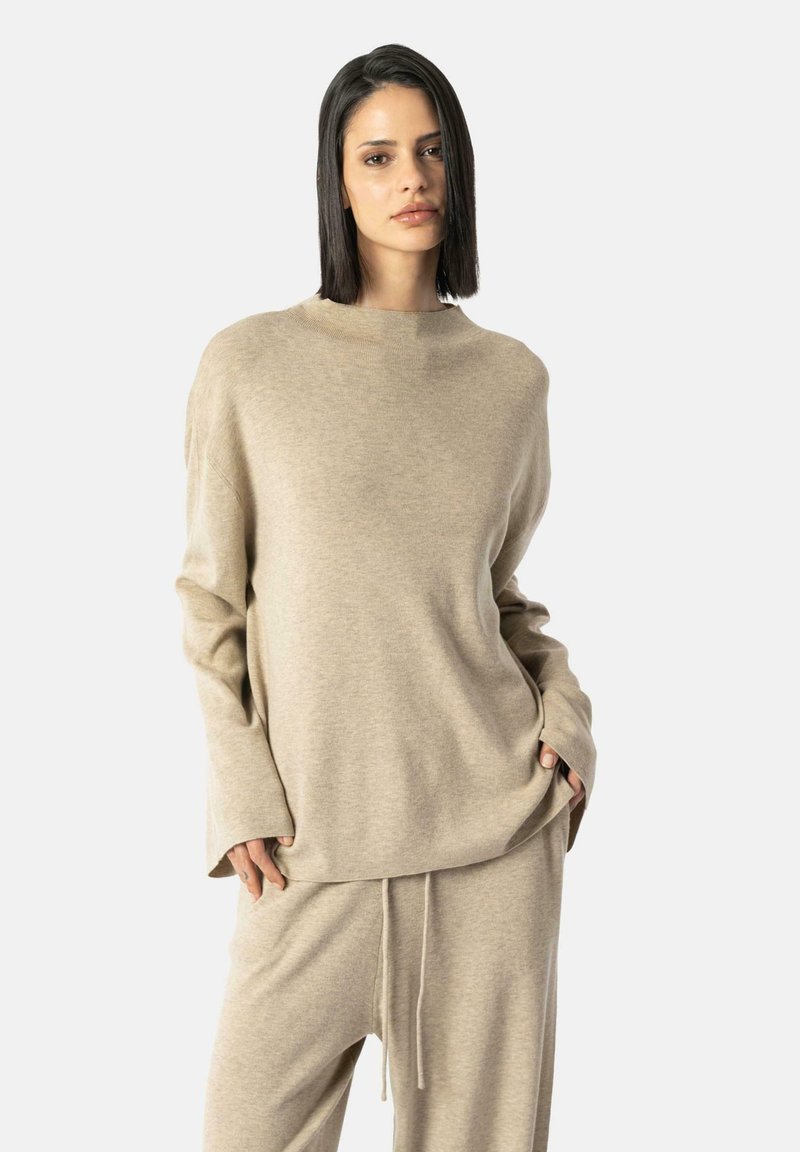 Beiger übergroßer Pullover mit hohem Kragen und langen Ärmeln, aus weichem Stoff. Kombiniert mit passenden, lässigen Hosen mit Kordelzug.