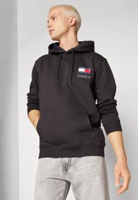 Sudadera negra de algodón suave con bolsillo canguro. Presenta un pequeño logotipo multicolor en el pecho y capucha con cordón.