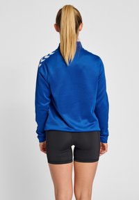 Blauwe lange mouwen top met een hoge kraag en witte chevronaccenten op de schouders, gecombineerd met zwarte aansluitende shorts. Gladde textuur overal.