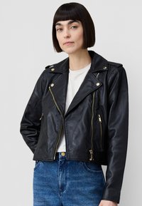 Svart läderjacka i bikerstil med guldfärgad metall, dragkedjeförsedda fickor och en hakkrage, bärs över en vit t-shirt och blå jeans.