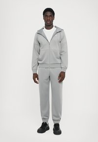 Lys grå tracksuit lavet af glat stof; lynlåsgennemdæk med hætte, ribbede ærmer, matchende bukser og sorte træningssko.