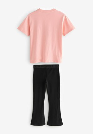 Világos rózsaszín pamut póló rövid ujjakkal, fekete harangszárú leggings-szel párosítva, amelyek márkás gumis derékrésszel rendelkeznek.