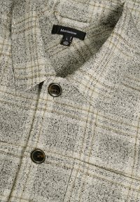 Cămașă gri deschis cu un model în carouri, având un etichetă neagră și un nasture închis la culoare. Material texturat cu accente galbene subtile.