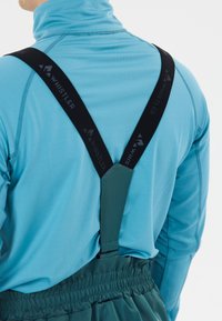 Mann in einem türkisfarbenen Langarmhemd und dunkeltürkisen Hosen mit schwarzen Hosenträgern, die auf seinem Rücken mit "Whistler" beschriftet sind.