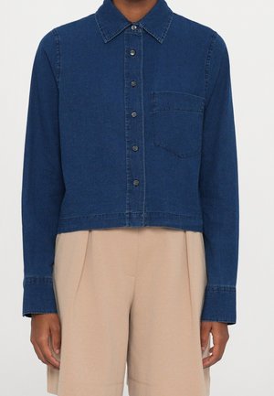 Personne portant une chemise en jean bleu foncé boutonnée avec une poche poitrine et un short beige plissé taille haute.