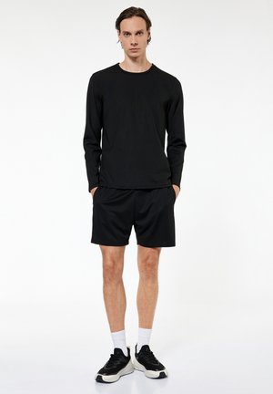 Mężczyzna stojący, ubran w czarną koszulkę z długim rękawem, czarne spodenki, białe skarpety do połowy łydki oraz czarno-białe trampki na białym tle.