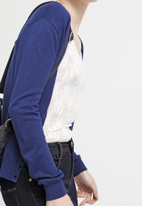 Námořnicky modrý svetr na knoflíky s dlouhými rukávy přes bílý krajkový top, doplněný tmavými džíny. Svetr má jednoduchý design a měkkou texturu.