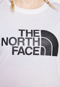 Bílý bavlněný tričko s velkým černým grafickým logem a nápisem "THE NORTH FACE" a zakřiveným designovým prvkem.