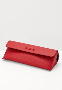 Raudona odiniais akinių dėžutė su tekstūruotu paviršiumi ir užlenkiamu dangteliu. Panašus įspaudžiamas "GUESS" logotipas priekyje.