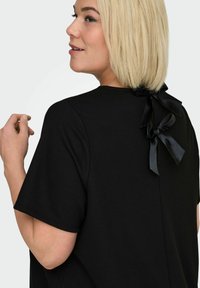 Czarny top z krótkim rękawem wykonany z teksturowanej tkaniny, z zapięciem z tyłu ozdobionym dwoma jedwabnymi kokardami. Minimalistyczny i elegancki design.