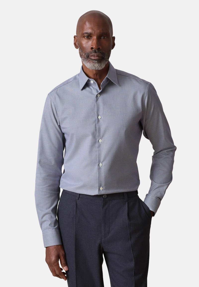 Uomo con barba grigia che indossa una camicia elegante a maniche lunghe a quadri azzurri infilata in pantaloni blu scuro, mano in tasca, sfondo neutro.