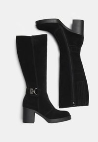 Bottes noires en cuir suédé jusqu'au genou avec un talon épais, dotées d'une fermeture éclair sur le côté et d'un détail de boucle en métal. Texture lisse avec un minimum de coutures.