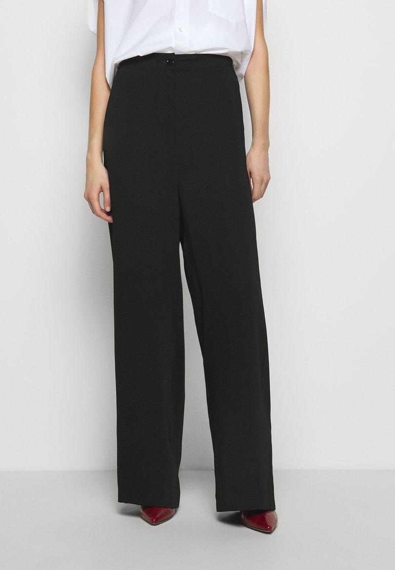 Svarta byxor med breda ben och en mjuk textur, hög midja och en enkel knappstängning. Bärs tillsammans med en ljus skjorta och röda klackar.