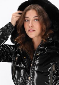 Jachetă puf neagră cu finisaj lucios, prevăzută cu glugă căptușită cu blană artificială, buzunare cu fermoar și accente din metal auriu.