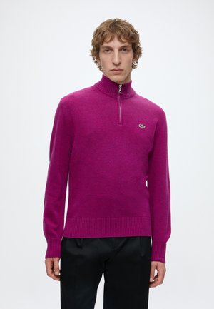 Jonge man die een felpaarse trui met een kwartritssluiting draagt met een klein groen krokodillenlogo op de borst en een zwarte broek.