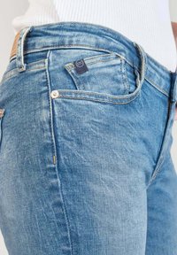 Jeans en denim bleu clair avec une surface texturée, dotés d'une poche avant, d'un bouton en métal et d'une étiquette logo sur la poche à monnaie.