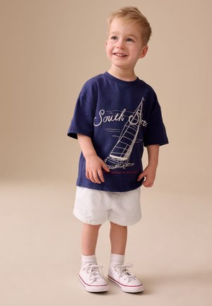 Niño pequeño sonriendo, con una camiseta azul marino con un diseño de velero, pantalones cortos blancos, calcetines blancos y zapatillas blancas, de pie sobre un fondo beige.