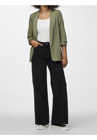 Blazer vert olive porté sur un débardeur blanc, associé à un jean noir taille haute à jambes larges et des baskets noires à semelles blanches.