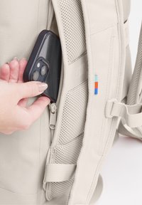 Zaino beige con superficie testurizzata; una mano che inserisce uno smartphone in una tasca laterale; cerniere visibili e piccoli accenti colorati.