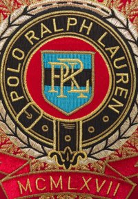 Blason brodé Polo Ralph Lauren avec lettrage doré, écusson bleu et noir, entouré de feuilles de laurier sur fond de tissu rouge.