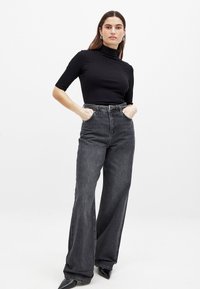 Melna ribota turtlenecka ar īsām piedurknēm, saskaņota ar augsta jostasvietas platām pelēkām džinsām. Džinsiem ir jostas ar sprādzēm un klasiskā džinsa tekstūra.