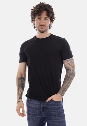 Uomo con capelli ricci e tatuaggi su entrambe le braccia, che indossa una maglietta nera e jeans blu, in piedi con una mano in tasca.