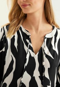 Žena se světlými hnědými vlasy, oblečená v topu s abstraktním vzorem v černobílé barvě, s výstřihem ve tvaru V a stahovacími šňůrkami.
